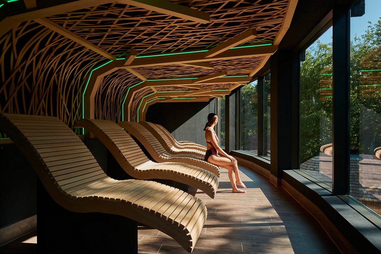 A woman sits on a curved wooden lounger, gazing outside; curved loungers line a spa relaxation room beneath a lattice ceiling with green lights and windows overlooking sunlit trees.